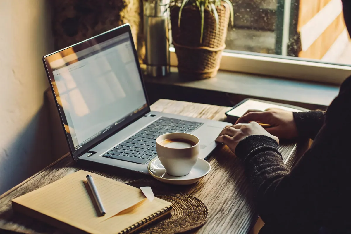 Persona escribiendo en un portátil junto a la ventana, con un café y una libreta